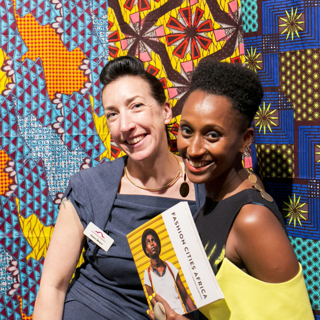 Curator Helen Mears and writer Hannah Azieb Pool pose with the Fashion Cities Africa book in front of brightly coloured Dutch wax print fabrics