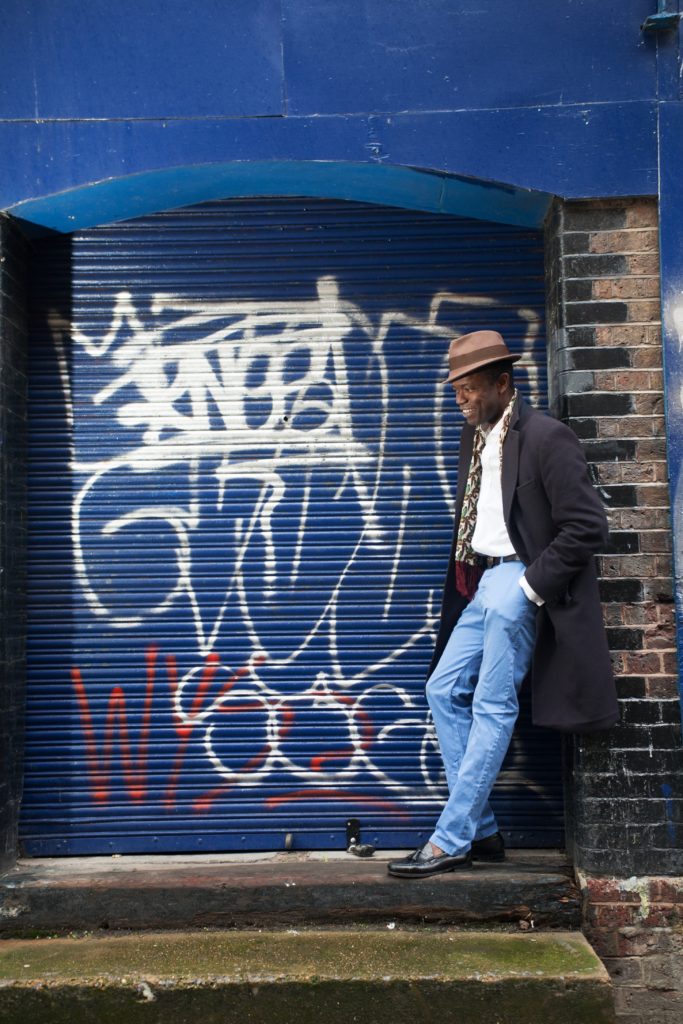 A man wearing a brown trilby, long black trench coat, white shirt and blue jeans stands in a blue doorway next to a shuttered shopfront