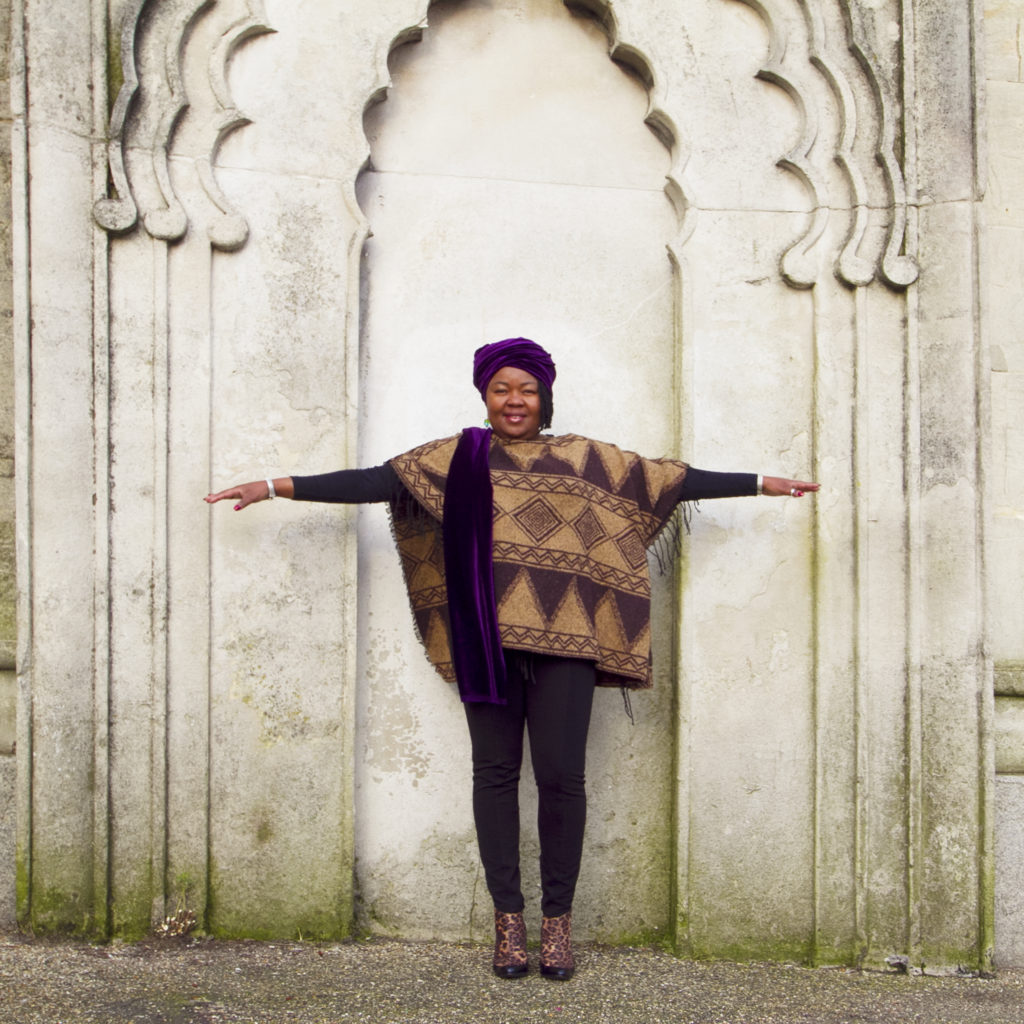 A woman stands with her arms outstretched in front of an Indian style arch on the Royal Pavilion estate, wearing a geometric print brown and black poncho over a black top and leggings, and a purple headwrap.