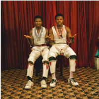 Colour photograph of two men seated in portrait studio, wearing white vest tops and white trousers, holding beadwork.
