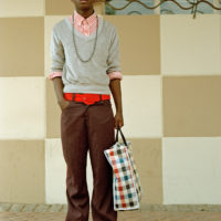 Colour photograph of a young man wearing brown trousers, a red striped shirt and grey jumper, accessorised with a grey hat, red sunglasses and belt, and a checked shopping bag