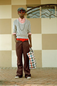 Colour photograph of a young man wearing brown trousers, a red striped shirt and grey jumper, accessorised with a grey hat, red sunglasses and belt, and a checked shopping bag