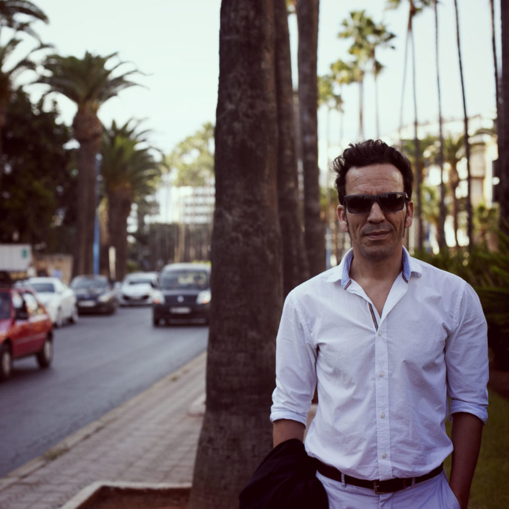 Said Mahrouf standing in a morroccan street with palm trees, wearing a white shirt and sunglasses