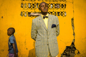 Colour photograph of a young man in a checked single breasted suit jacket with his hands behind his back, standing in front ot a yellow wall. A young boy in a blue T shirt is walking past on the left of the frame.
