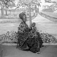 Studio portrait of a woman wearing voluminous robes, headwrap and flipflops, with a small child in her lap.