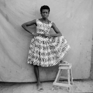 Portrait of a woman in a sleeveless, knee-length print dress with a full skirt, posing with her hands at her waist and one leg on a stool, in front of a cloth backdrop.