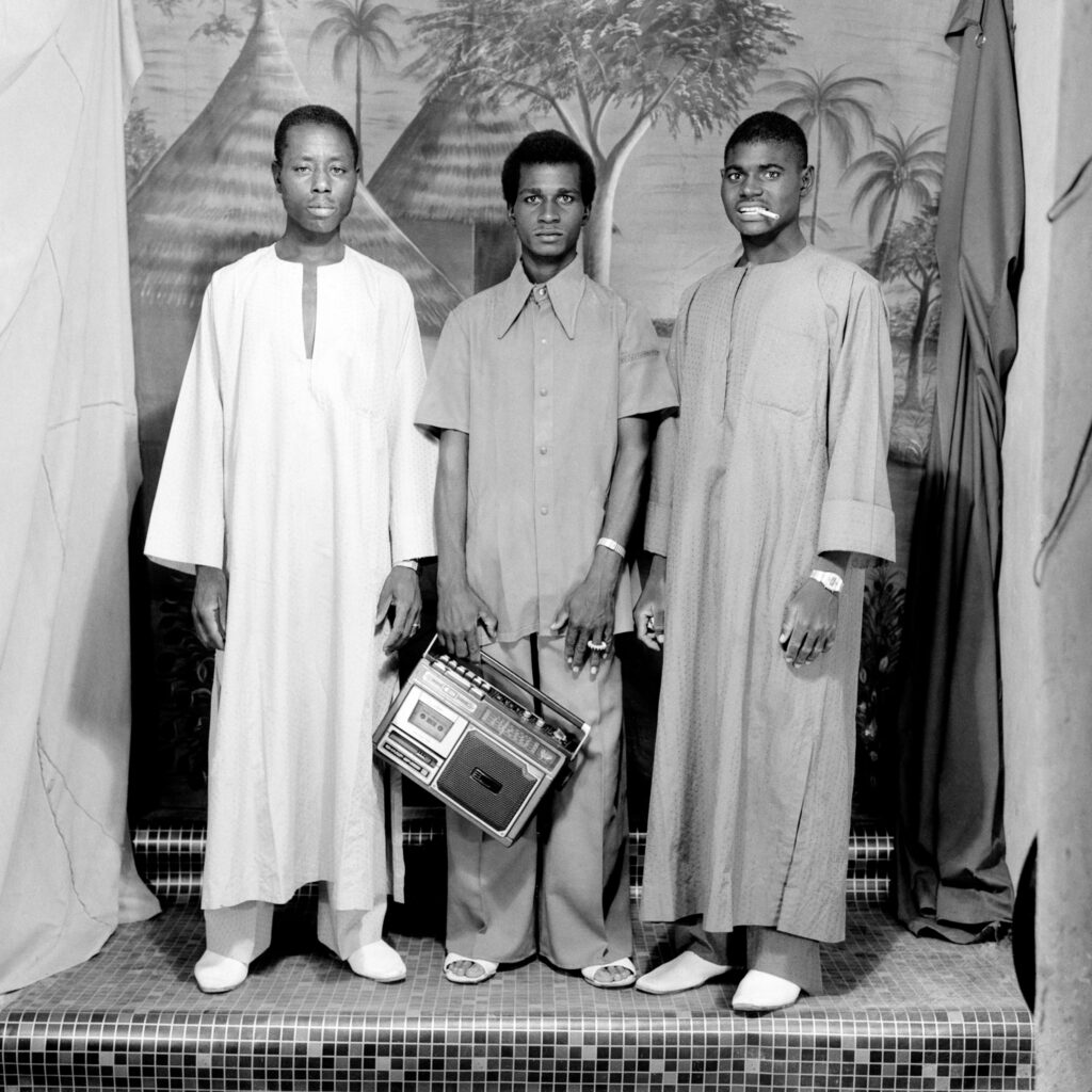Studio portrait of three men standing in front of a painted cloth backdrop, one holding a portable stereo.