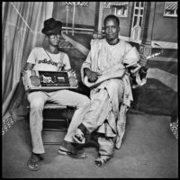 Studio portrait of two young men, one wearing an Adidas T shirt and black hat, holding a portable stereo on his lap. The other man is wearing a long robe and has a stringed musical instrument.