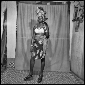 Studio portrait of a young woman wearing a white bra, wrap skirt, scarf tied around her waist, an elaborate headwrap and platform shoes.