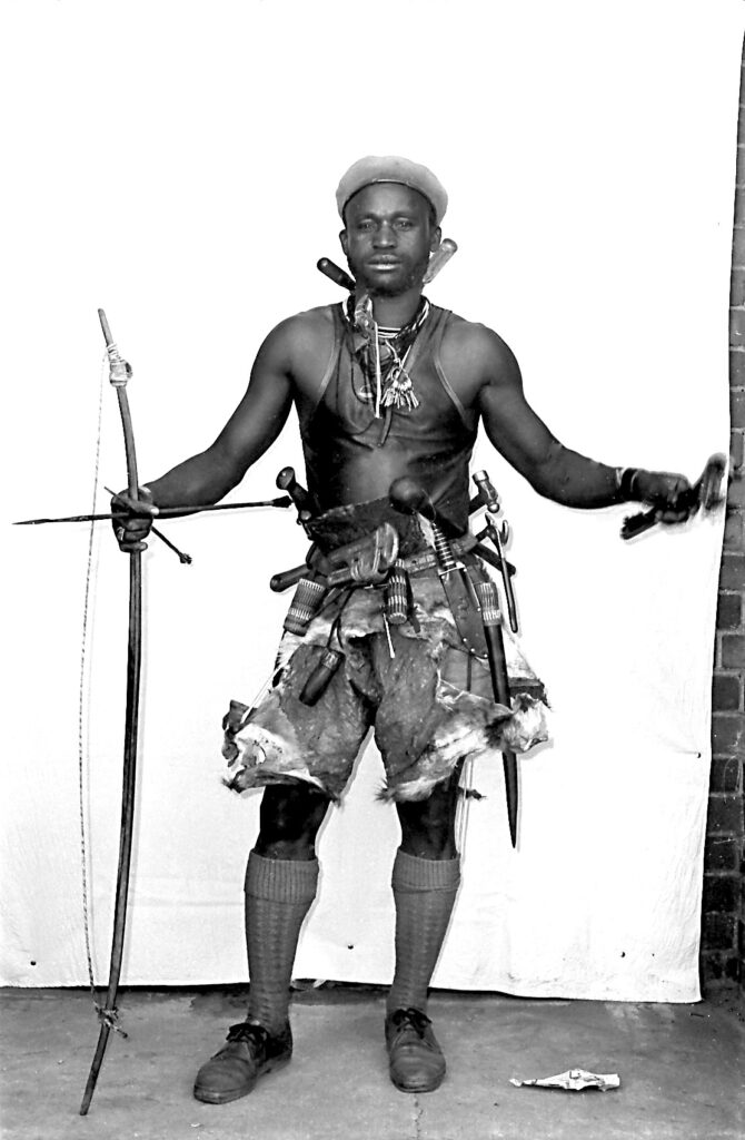Black and white photograph of man in vest and shorts with hunting gear and bow and arrow