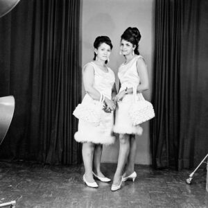 Black and white Studio portrait of two young women women wearing matching white dresses, shoes and handbags.